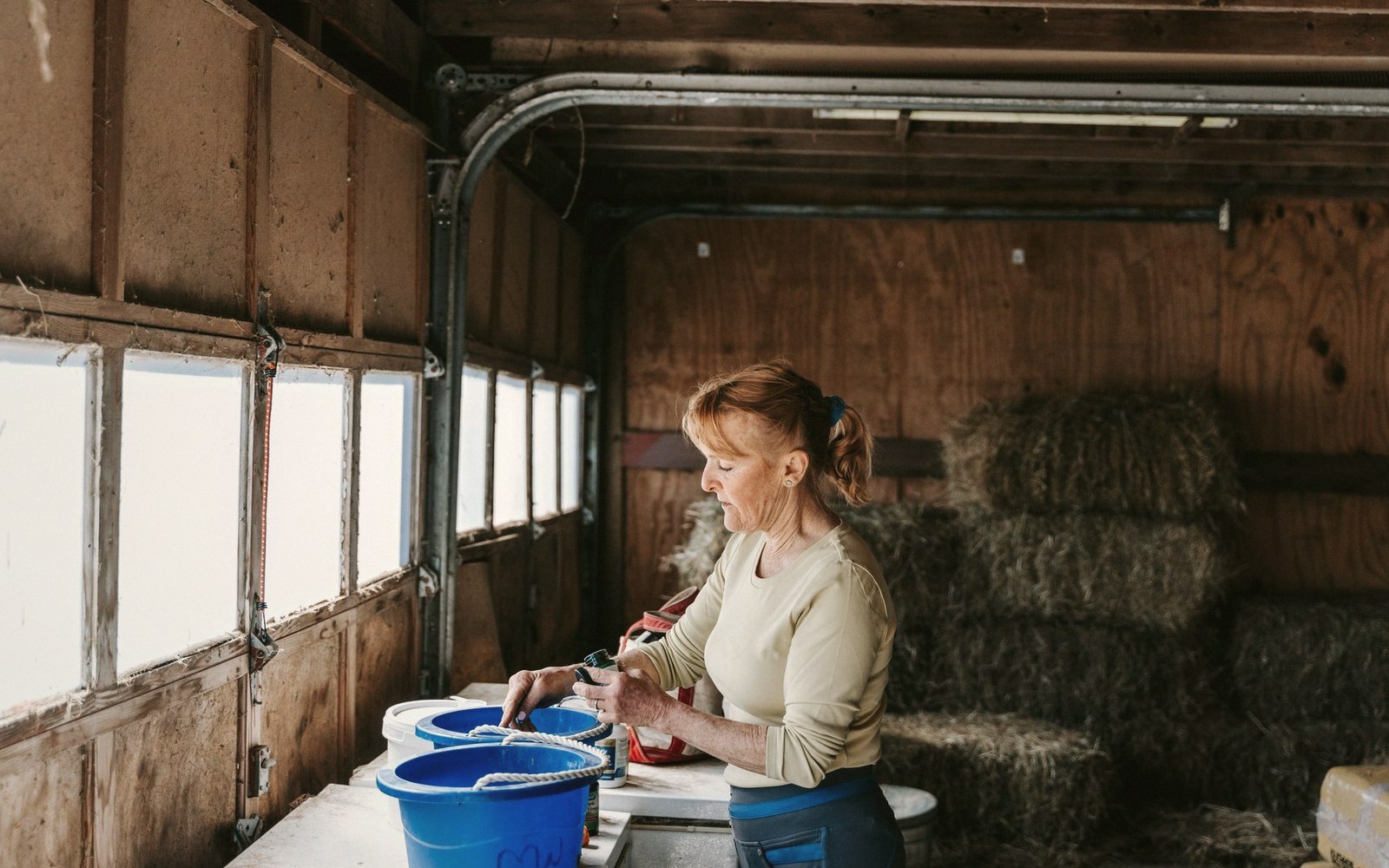 A woman ties rope to a bucket in a barn stacked with hay bales