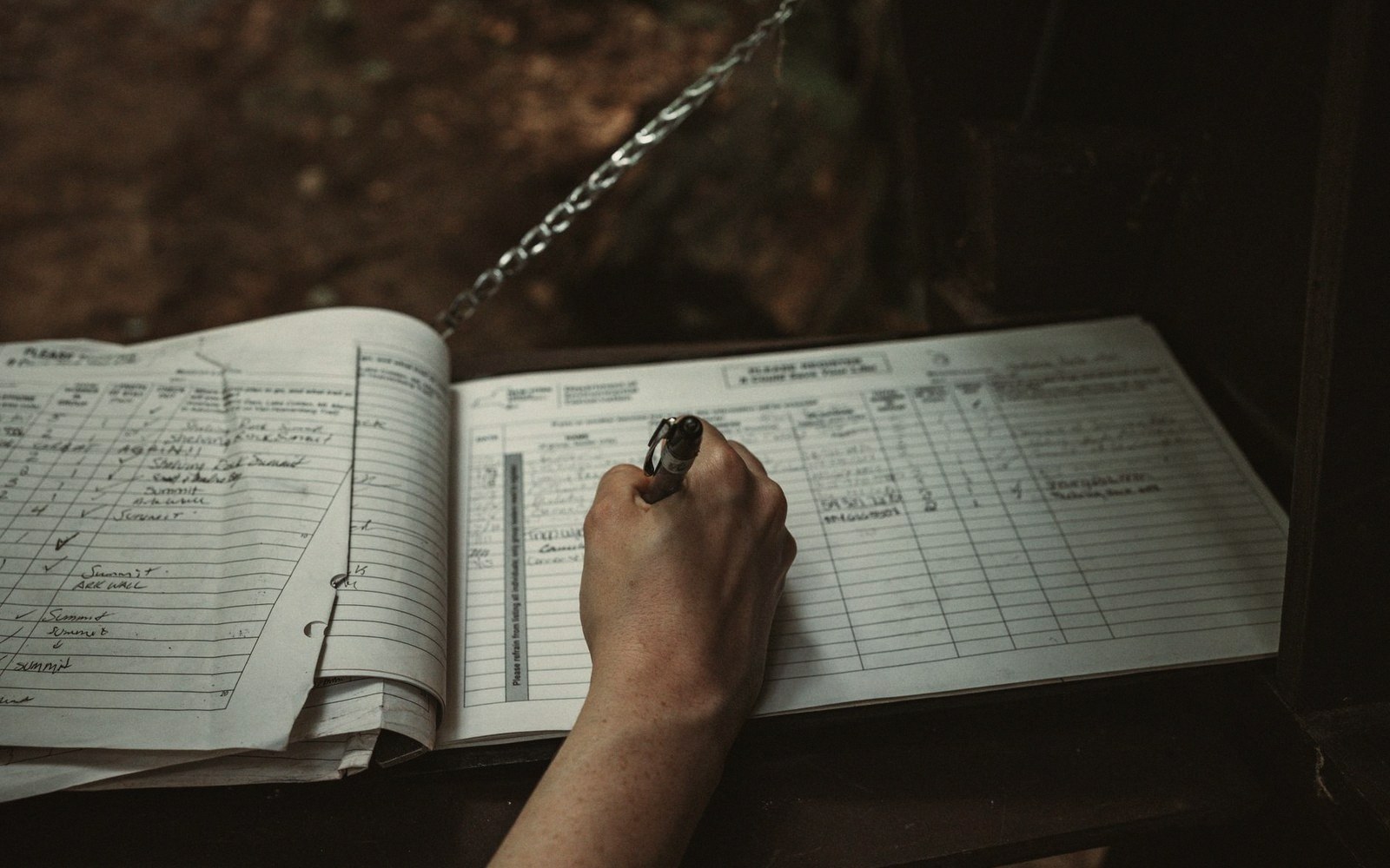 A hand fills in entries on a lined visitor logbook attached by a chain